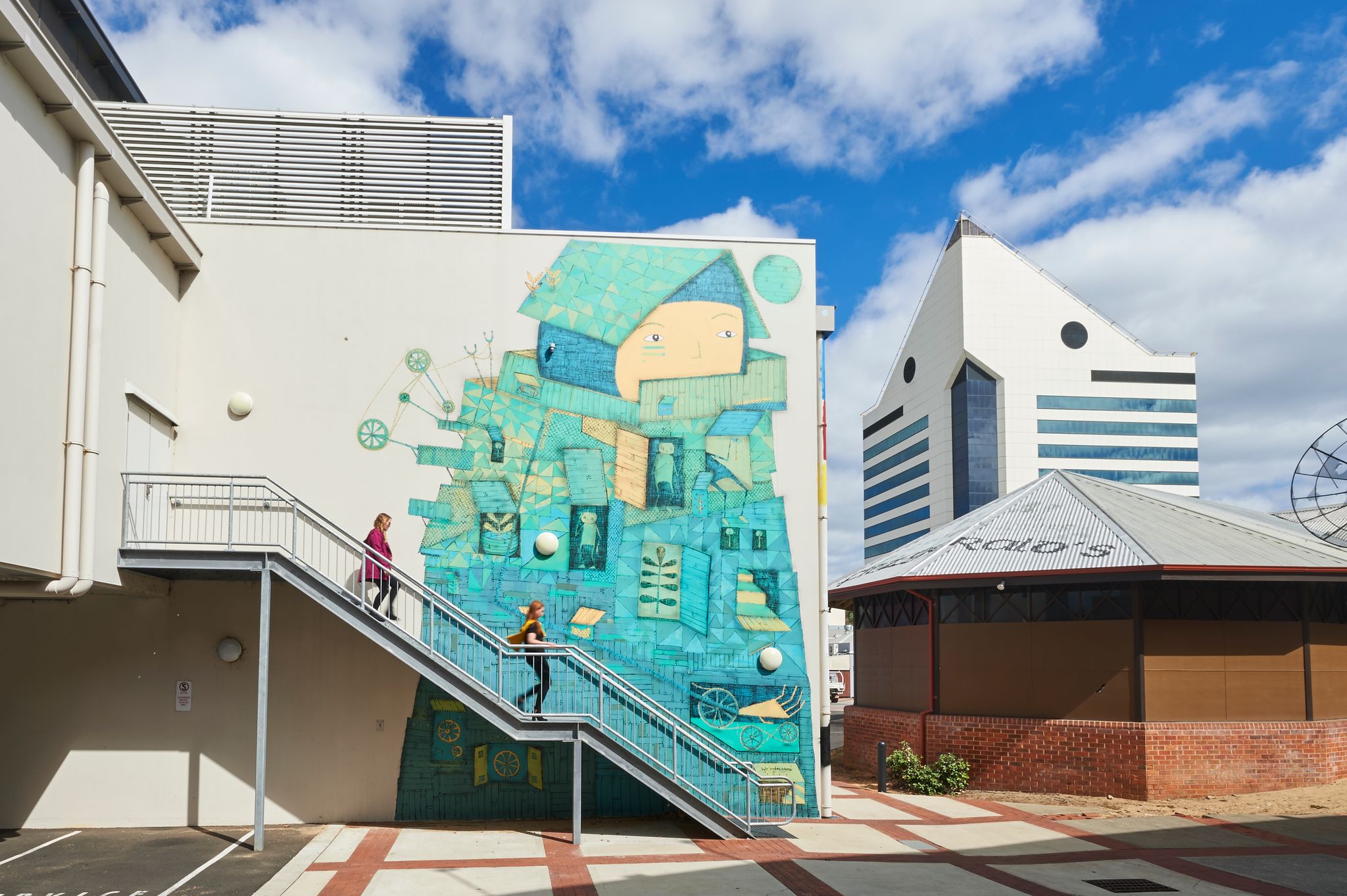 2 women walk down a steel staircase in front of a green mural of a face