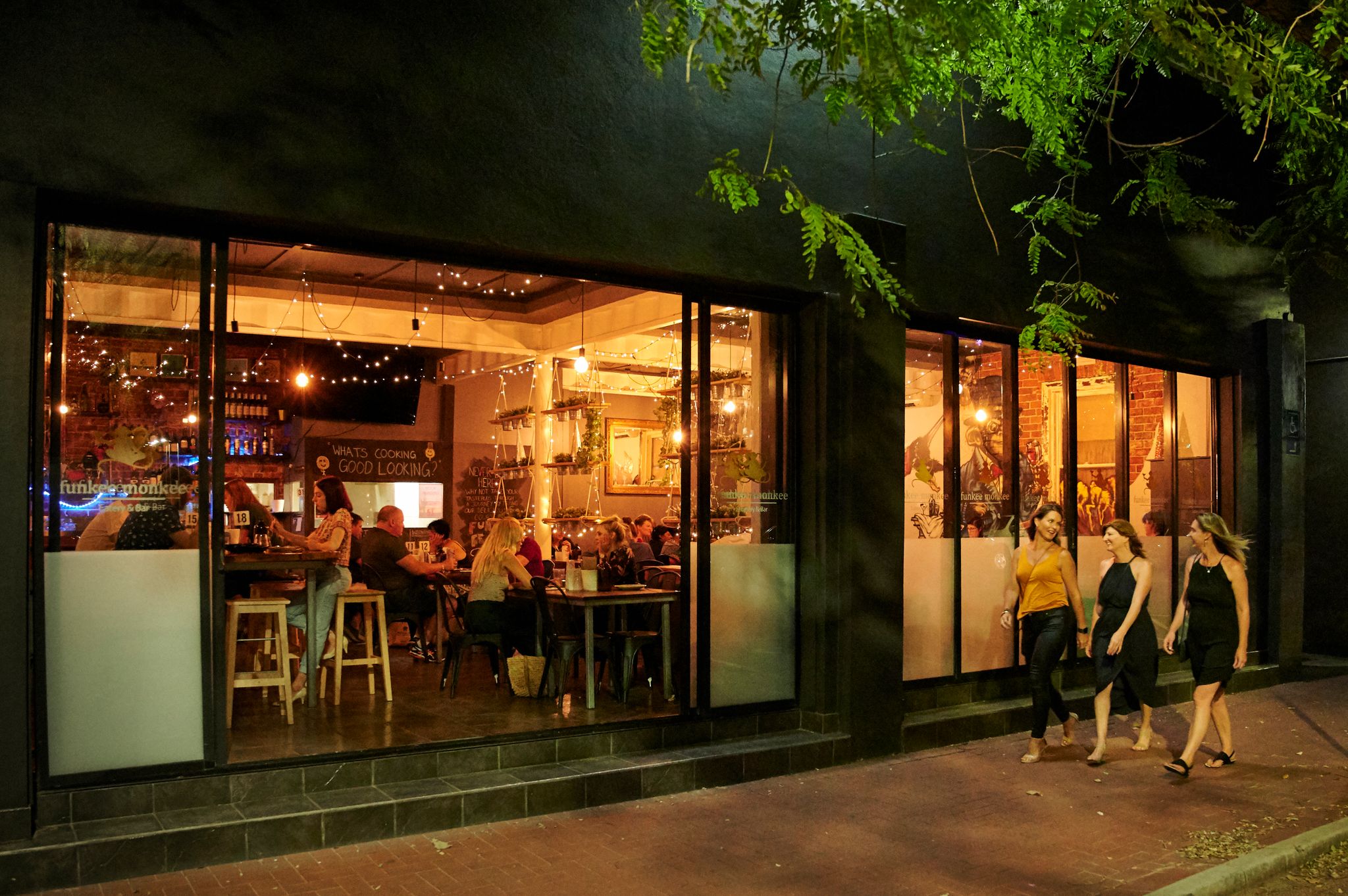 Three women walk past a bustling restaurant in Bunbury filled with fairy lights