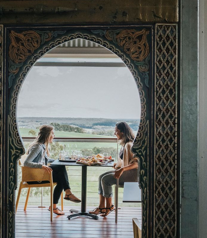 Two women sitting on a wooden deck behind green door having wine and food from a platter