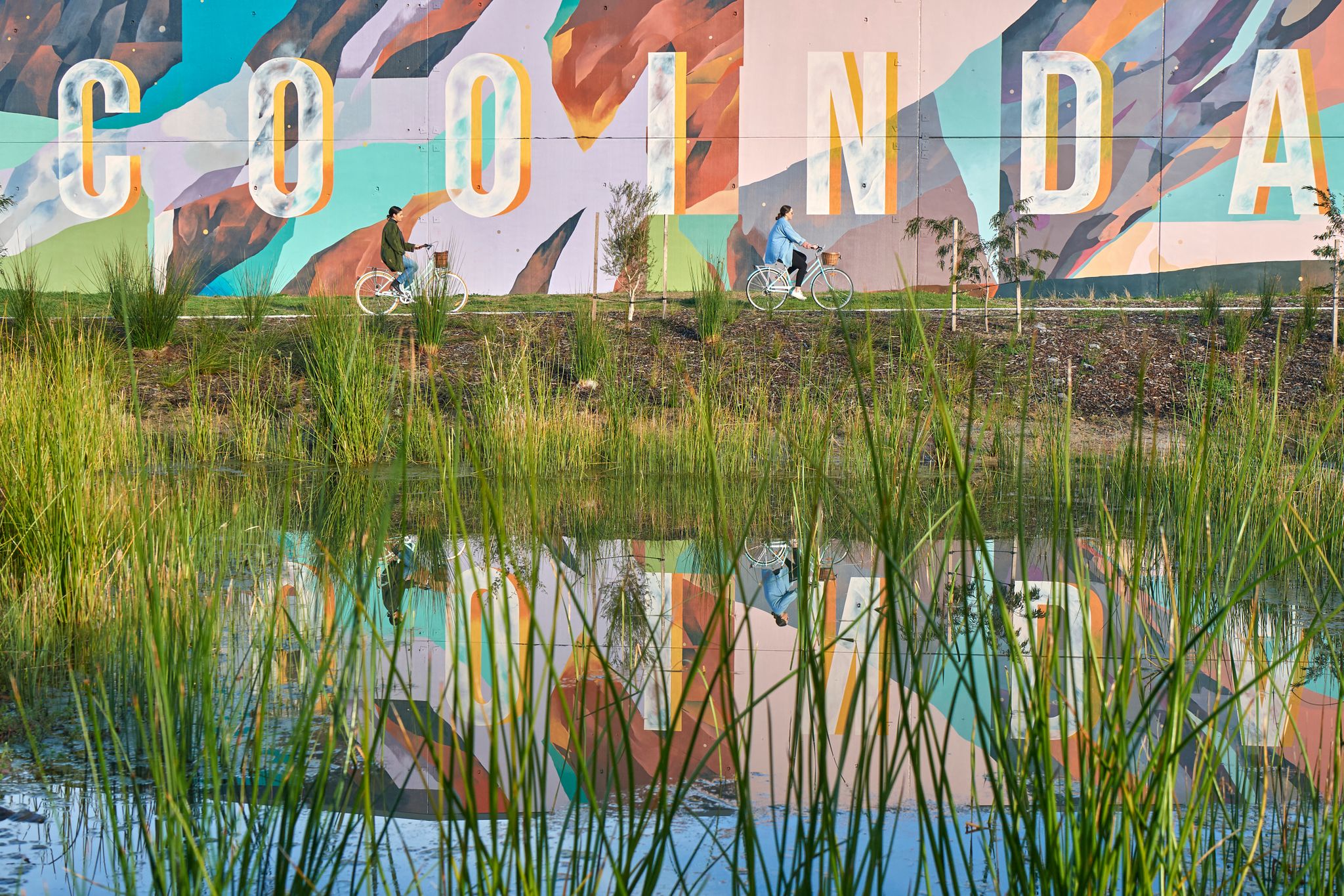 2 women on bikes riding part a painted wall with the words Cooinda on it. 