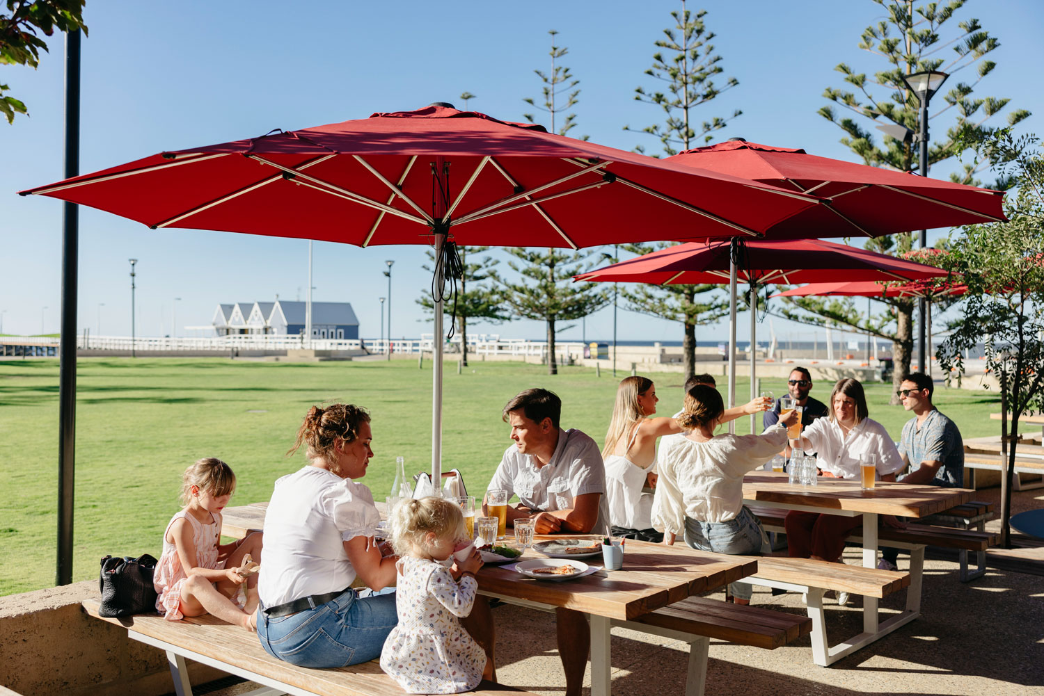 A family sits under an umbrella at the table in a brewery with a foreshore and ocean in the background. 