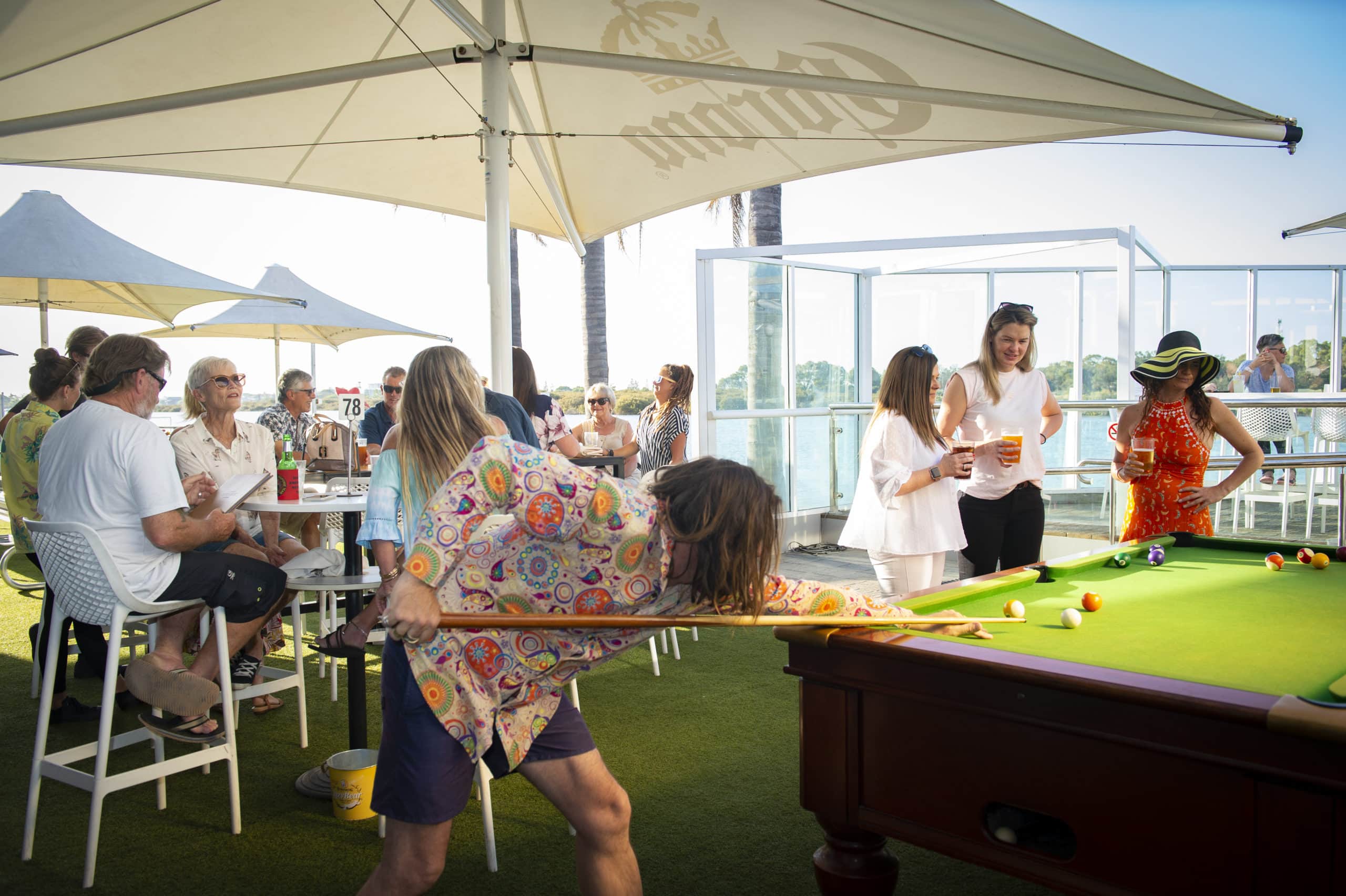 A man playing pool amongst other patrons dining on a deck on the water